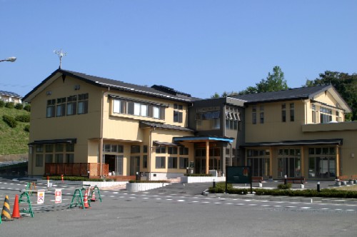 淡い黄色の外壁と黒い屋根が特徴的な2階建ての建物で、広いアスファルトの駐車場が手前に見える、歌津公民館の外観写真