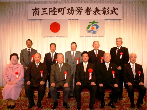 「南三陸町功労者表彰式」と書かれた横断幕が掲げられた会場で、町長と受賞者が2列に並び、胸に赤いリボンを付けて記念撮影に応じている様子の写真