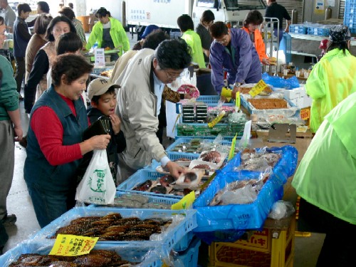市場で、多くの人々が、青いプラスチックのケースに並べられた魚介類や加工品を見ている産業フェアの様子の写真