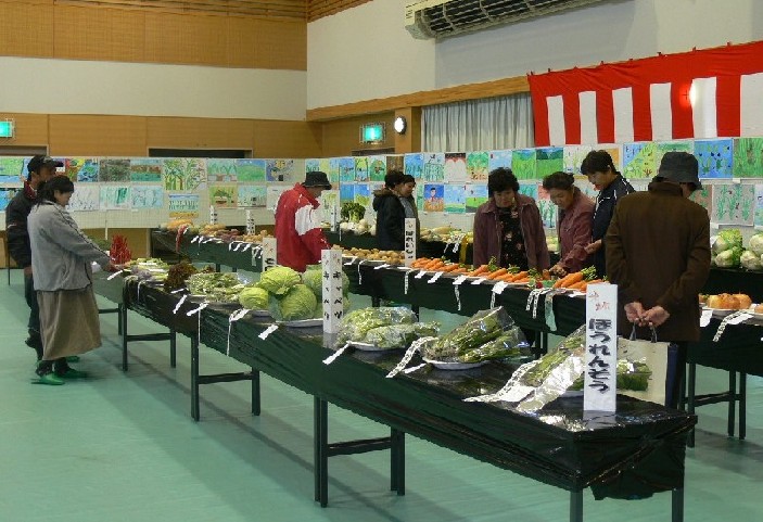 広々とした屋内のイベント会場で、人々が長いテーブルに並べられたキャベツやニンジンなどの新鮮な野菜を品定めしている南三陸町産業フェアの様子の写真