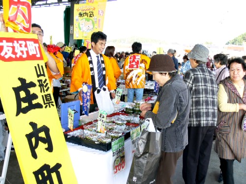 屋外で開催されたイベントで、オレンジ色のはっぴを着た関係者が、商品を勧めるように笑顔で女性客の対応をしている、活気ある販売ブースの様子の写真