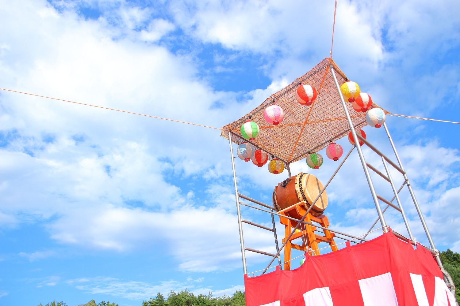 青空を背景に、赤と白の幕で囲まれた櫓の上に設置された大きな和太鼓と、その周囲に吊るされたカラフルな提灯が見える写真
