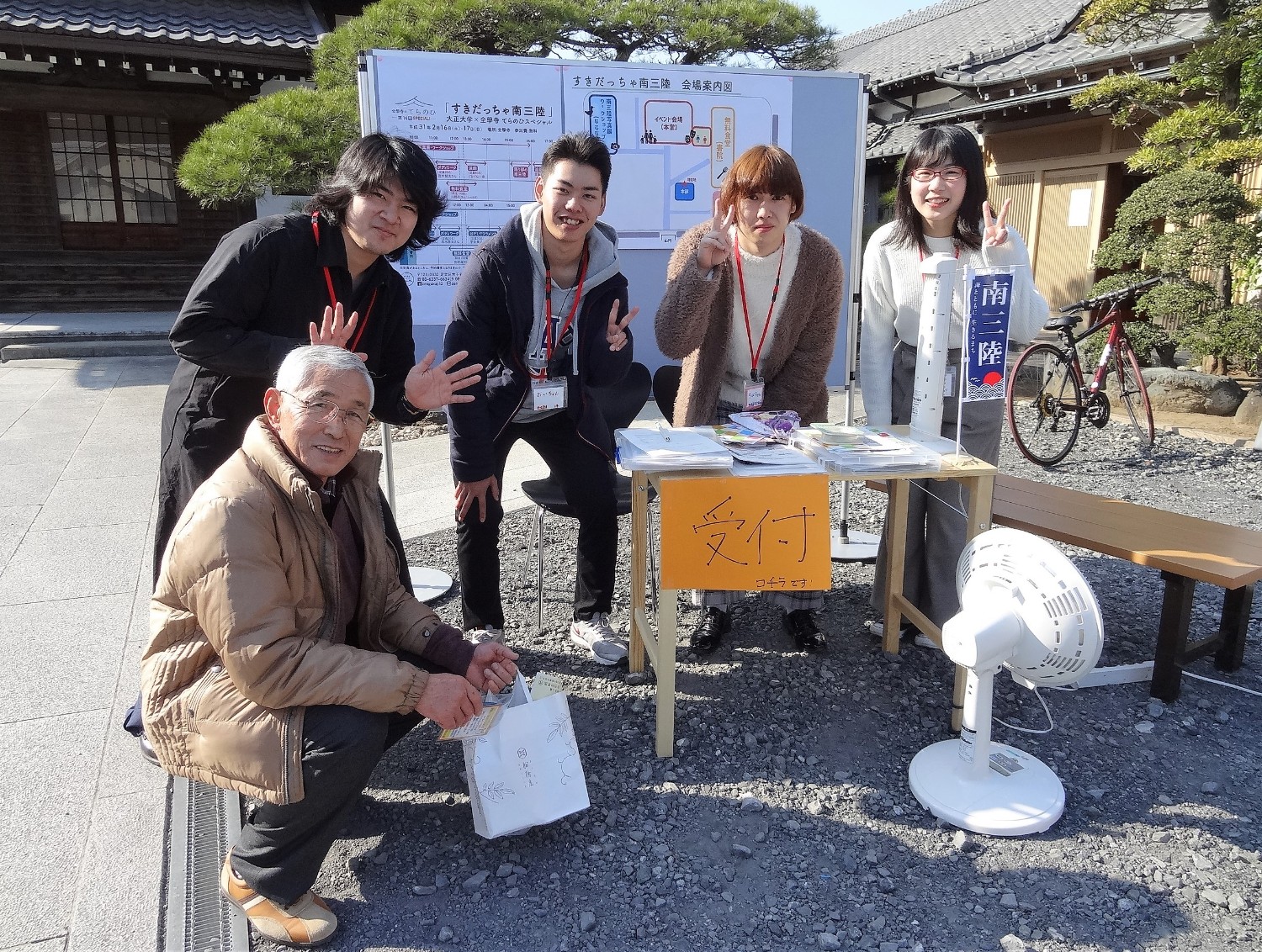 屋外の受付テーブルの前に5人の人物が集まり、カメラに向かってピースサインをしている写真