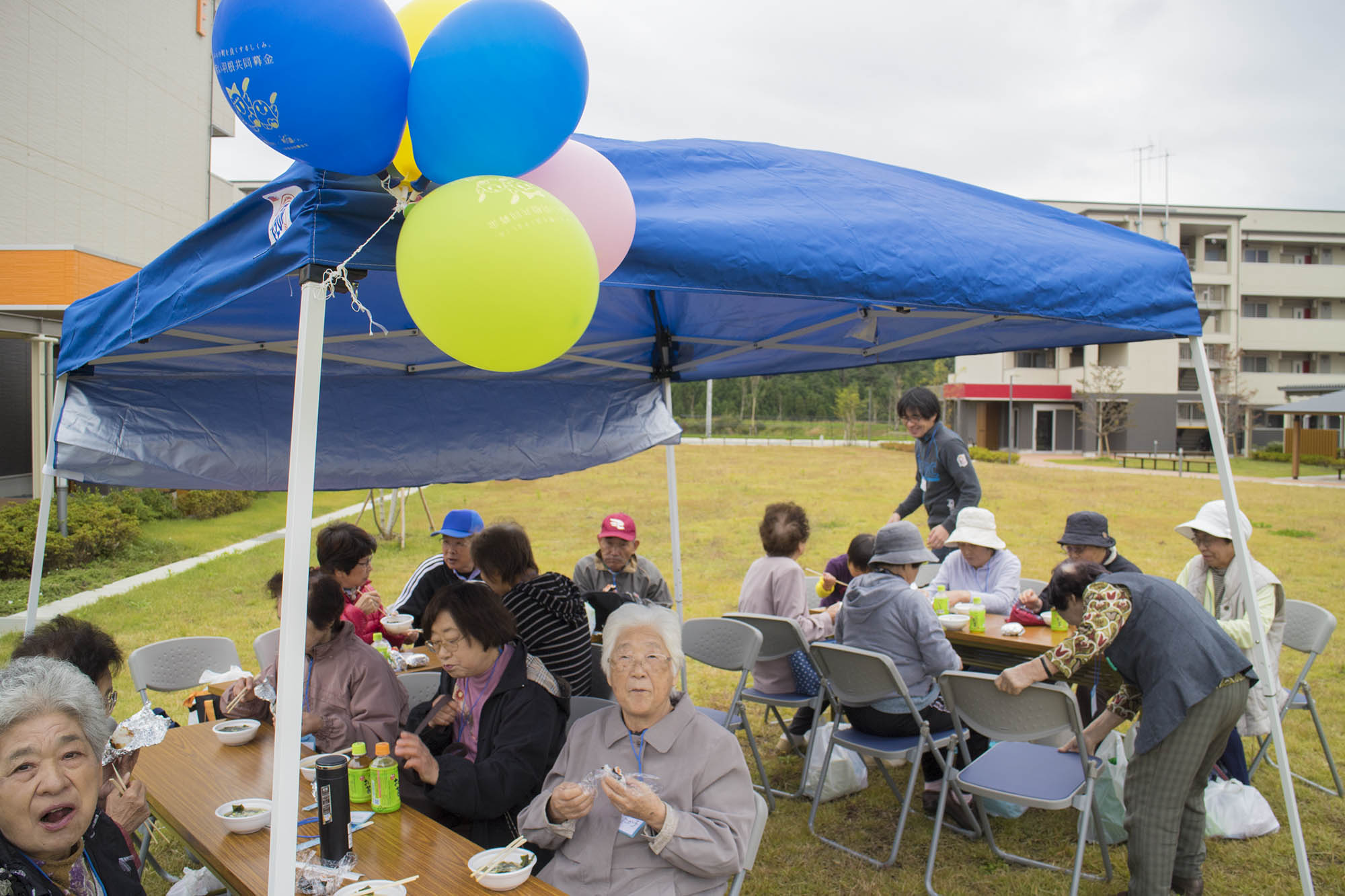 青いテントの下で多くの人々がテーブルを囲んでおにぎりや汁を食べて談笑しており、テントには風船が結びつけられている地域の運動会の写真