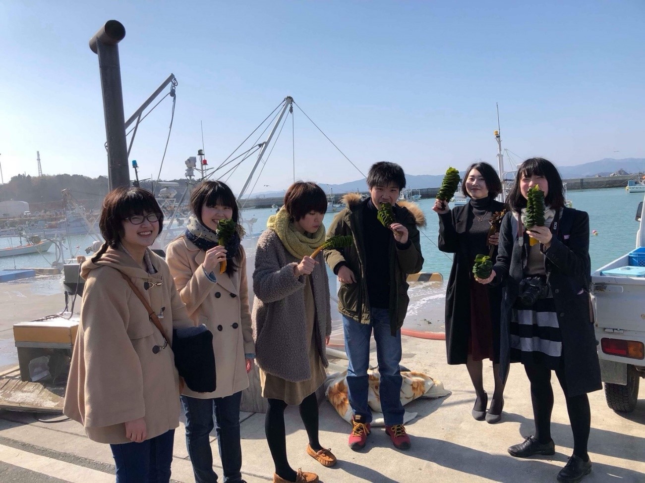 漁船が見える港で、6人の若者たちが楽しそうに海藻を手に持ち、口に持っていったりして笑顔を見せている写真
