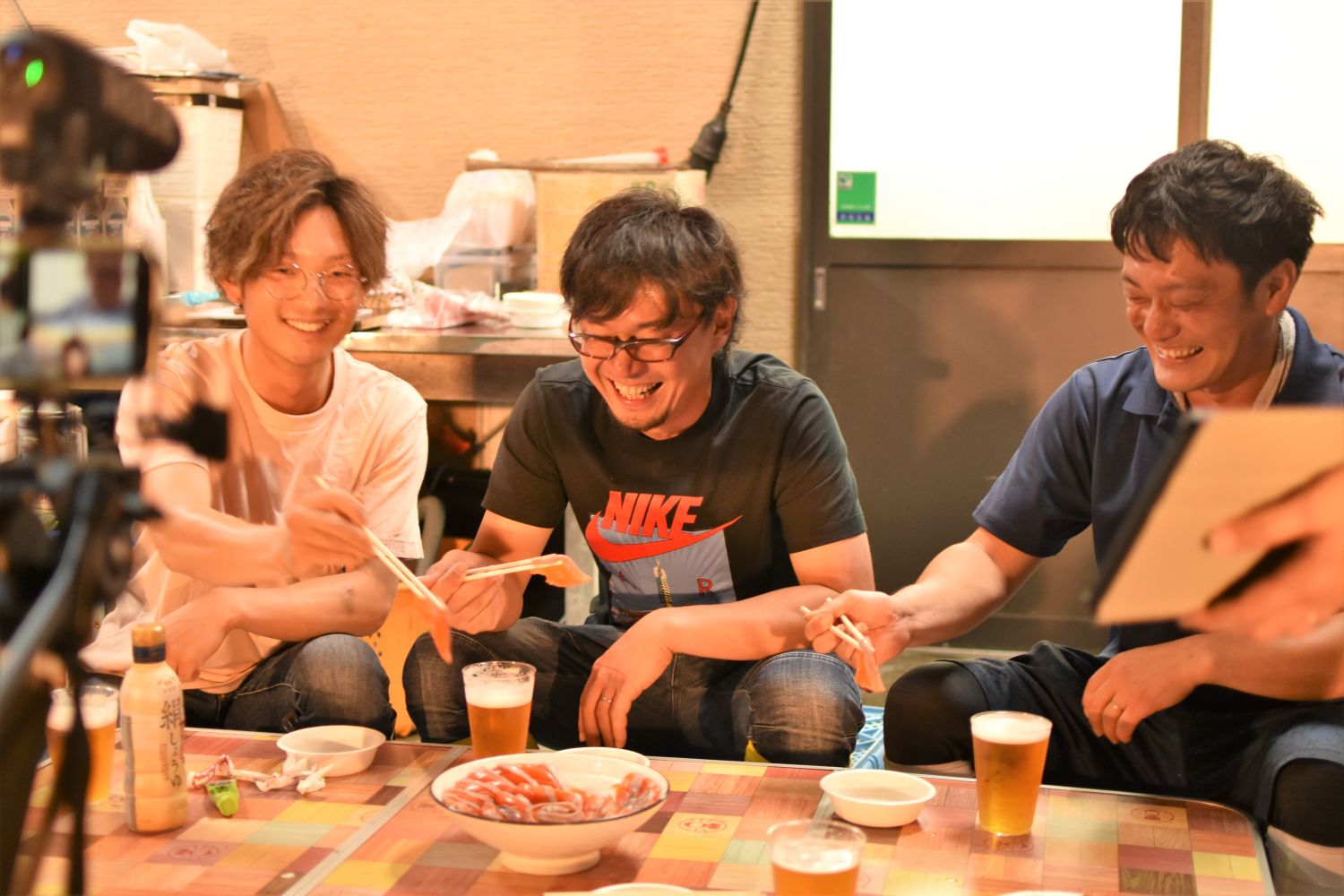 三人の男性が笑顔で刺身を箸で取りながら楽しそうにビールを飲んでいる様子を、カメラで撮影している写真