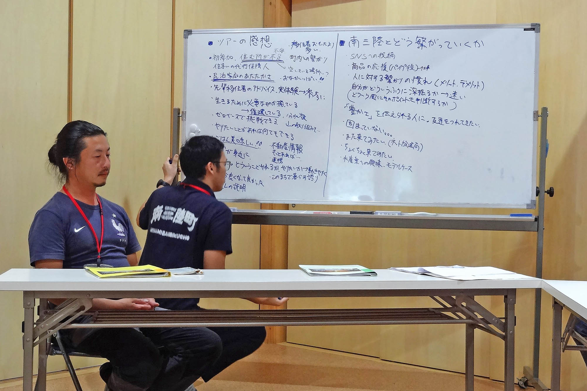 木の内装の会議室で、男性2人がホワイトボードの前に座り、ホワイトボードにはツアーの感想や地域振興の議論内容が書かれている写真