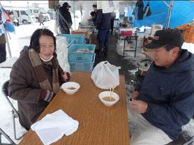 汁が入ったお椀や購入品が入ったレジ袋が置かれたテーブルの両側に防寒着を着た男女が笑顔で向かい合って座っている写真