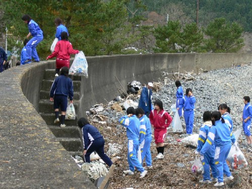 ジャージを着た多数の学生たちが、ゴミ袋を手に持って海岸沿いのコンクリートの護岸の下で、瓦礫やゴミを熱心に清掃している様子の写真