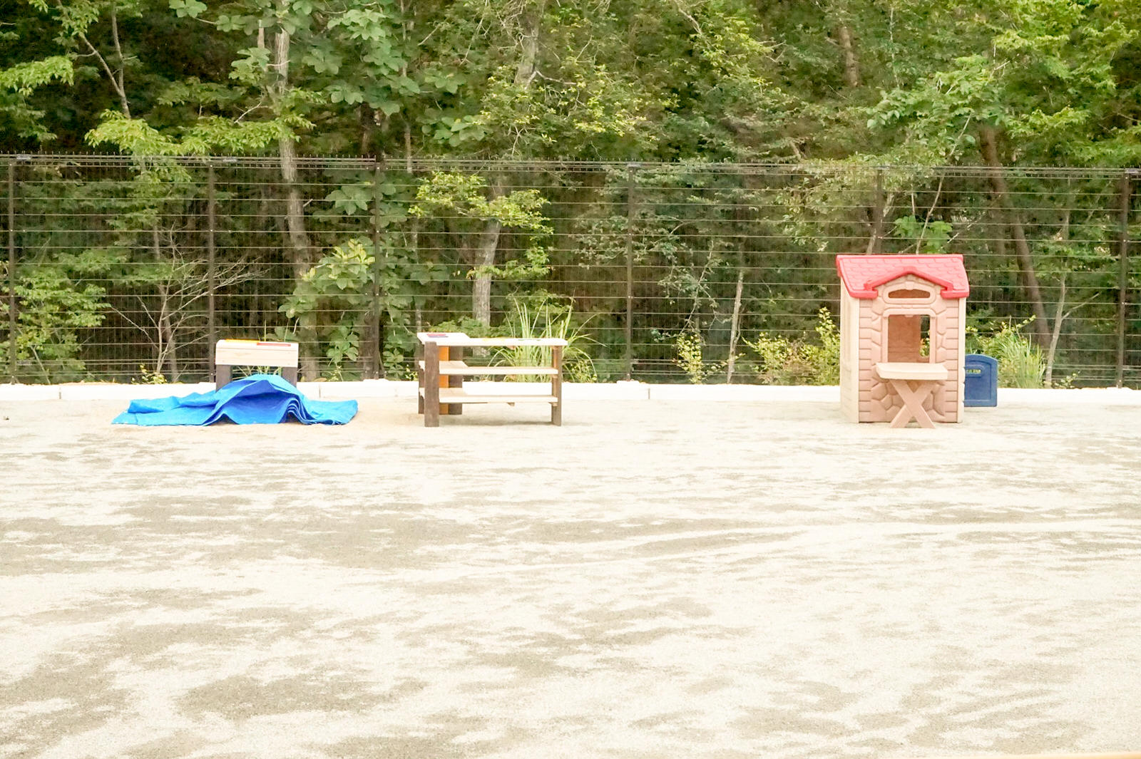 広い砂場に棚や赤い屋根のプラスチック製の遊び小屋があり、奥のフェンスの裏には緑の木々の林が広がっている写真