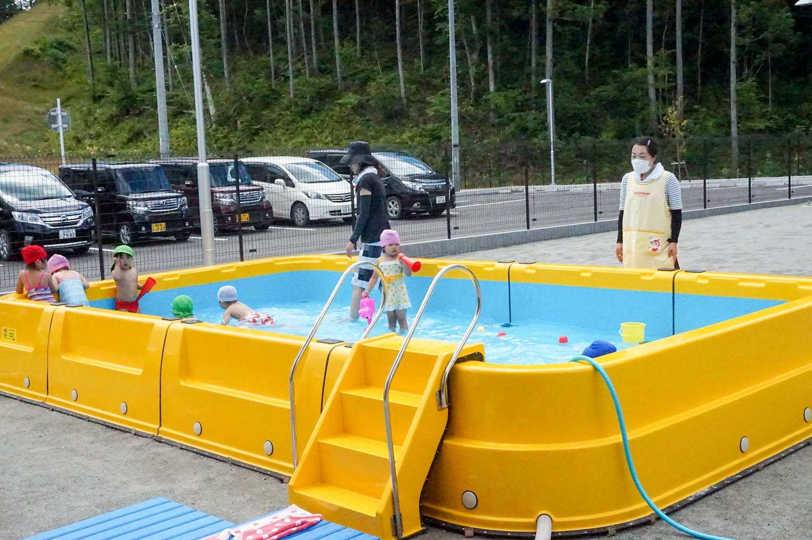 駐車場横にある、フェンスで囲まれた園庭に、階段付きの長方形の黄色い小型プールが設置され、中で子どもたちが水遊びをしており、傍で2人の先生が子どもたちを見守っている写真