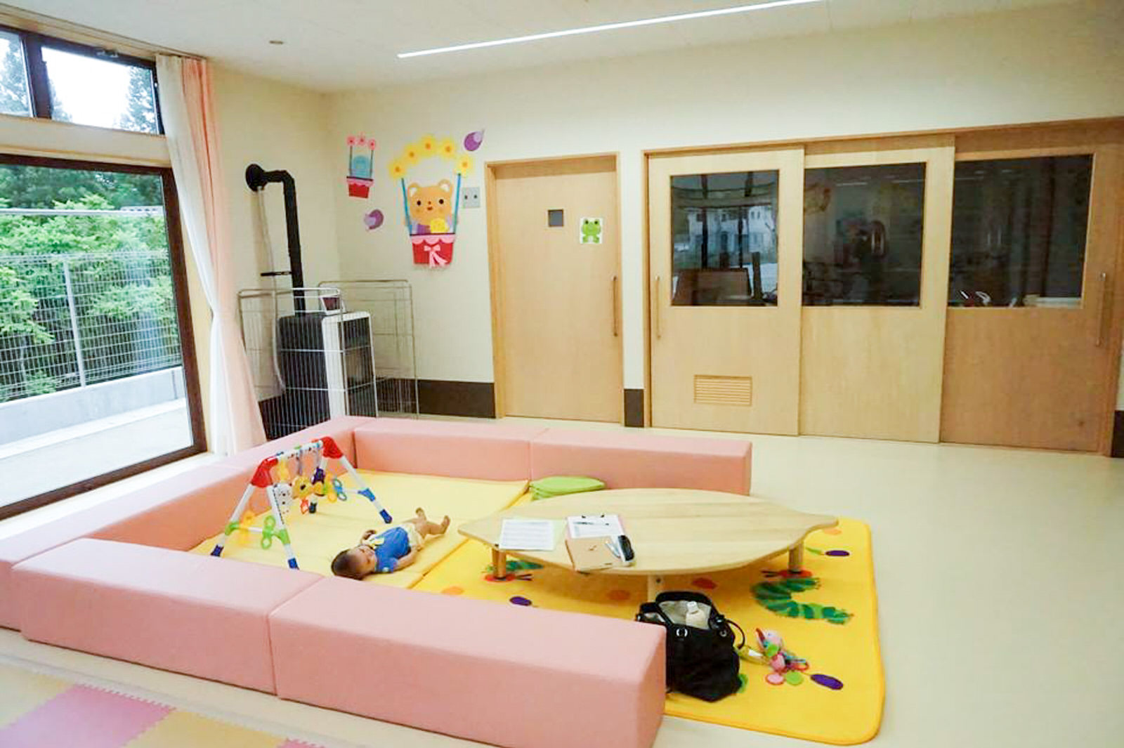 左側の大きな窓から自然光が差し込み、奥の壁には複数の木製扉が並んでいる室内の遊び場に、ピンク色のクッションで囲まれ黄色のマットが敷かれたスペースがあり、そこに小さな木製テーブル、幼児用の遊具が置かれ、傍に青いシャツを着た赤ちゃんが寝転んでいる写真