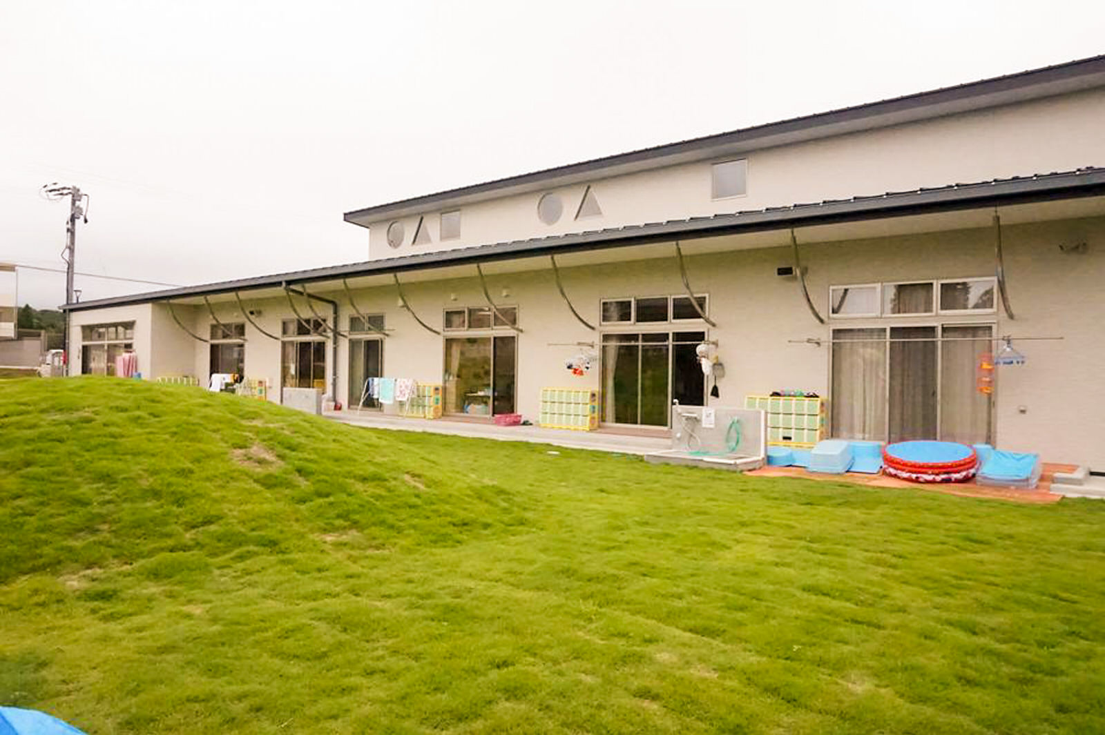 大きな窓が並ぶベージュの外壁の建物の前に芝生の園庭があり、小さな芝生の丘がある写真