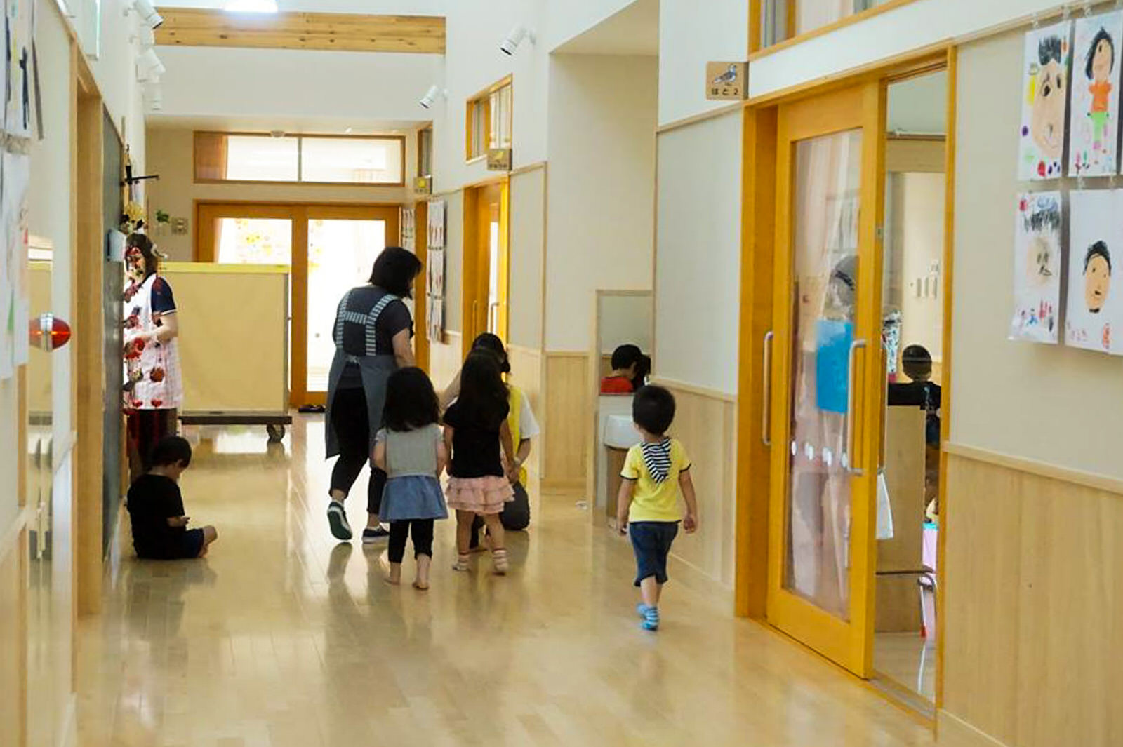 壁には子どもたちの絵が飾られ、右側には別室へ続く引き戸があり、奥の窓から自然光が差し込む明るい木目調の床が広がる廊下には園児の絵が飾られ、歩いたり床に座ったりしている複数の子どもたちと2人の保育士がいる保育所内の写真