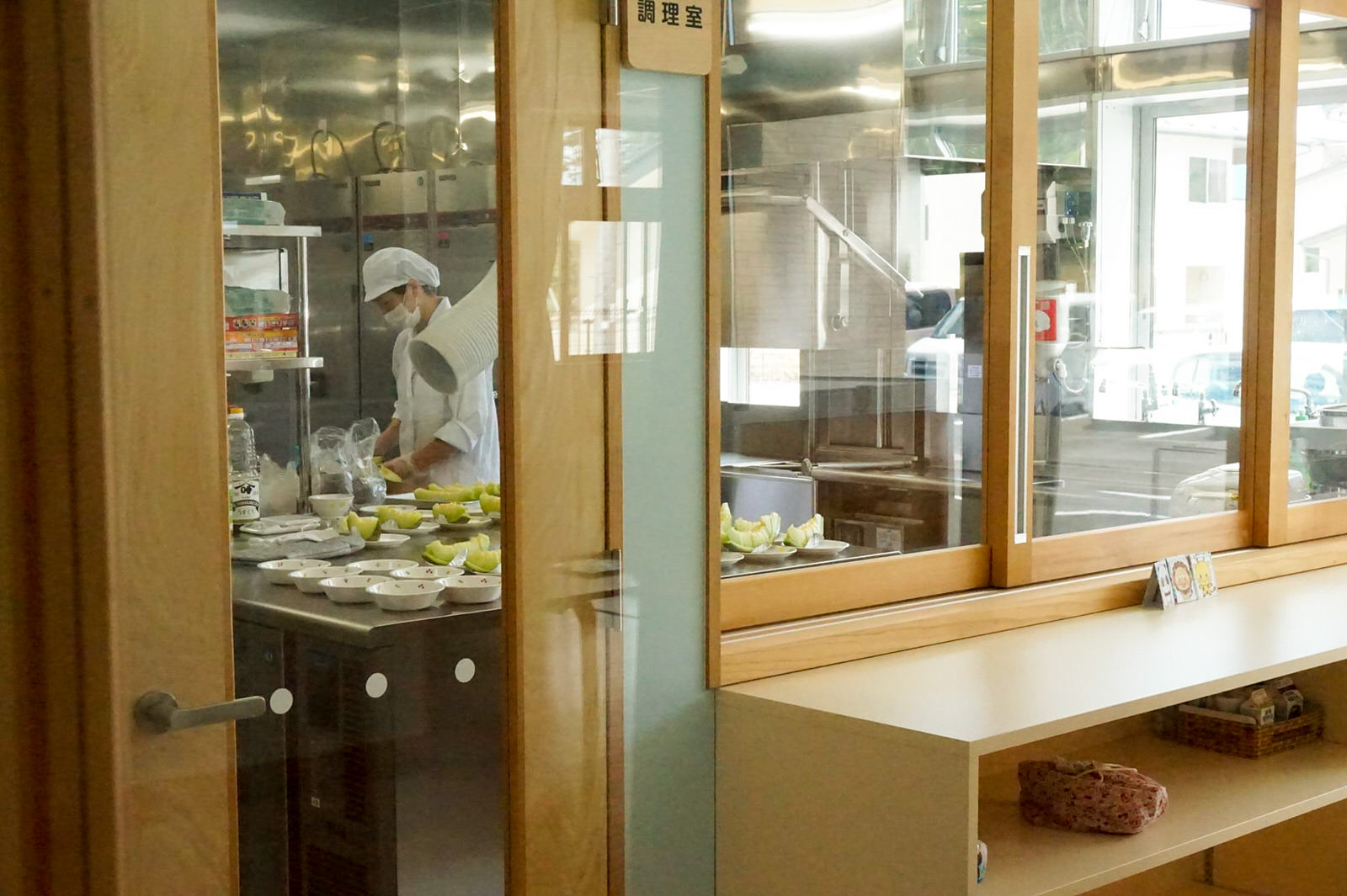 ガラス窓と扉越しに見える調理室で、白い制服と帽子を着用した調理師が複数の皿やボウルを並べた調理台で料理を準備している写真