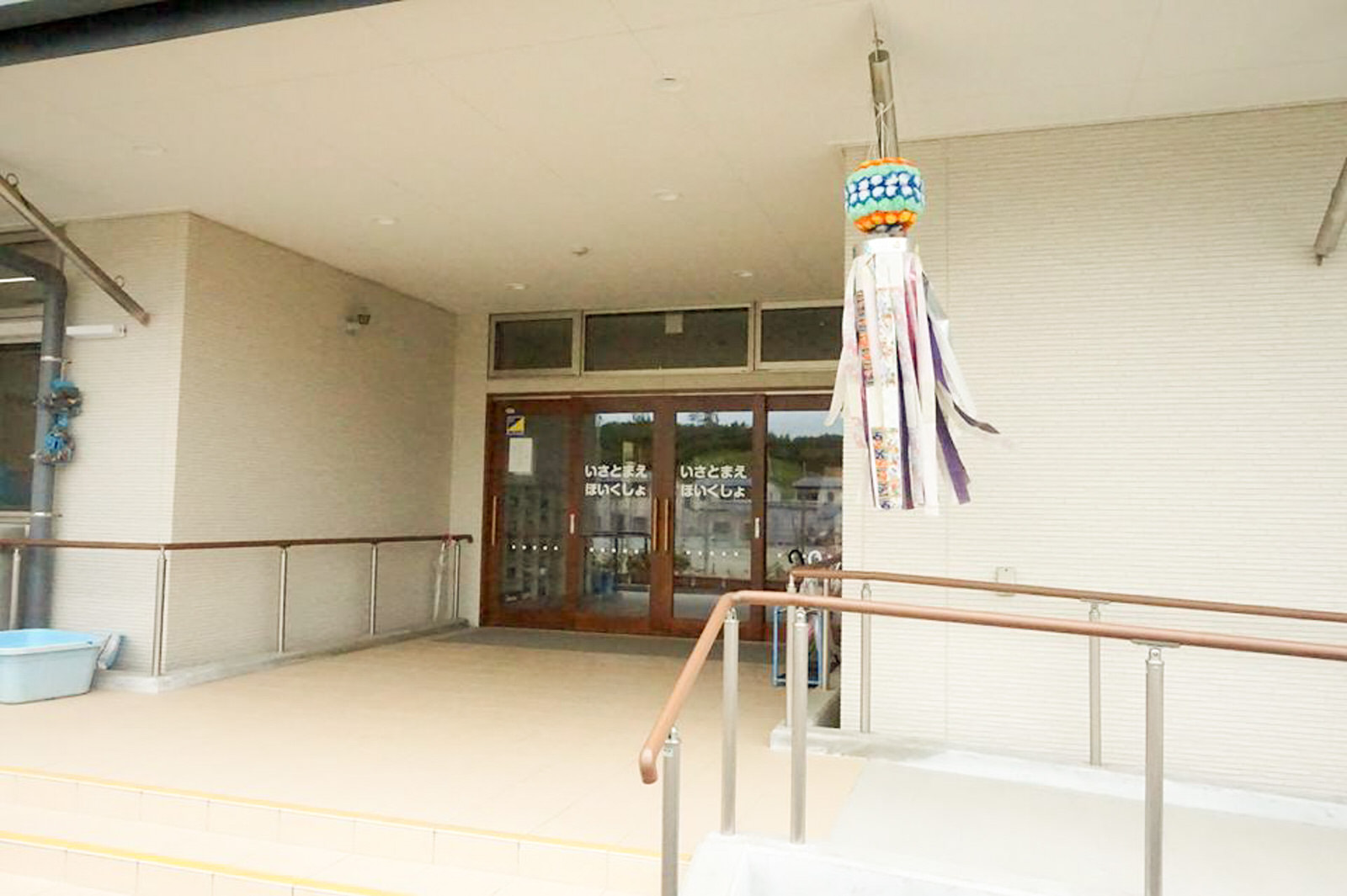 「いさとまえ ほいくしょ」と書かれた正面のガラスの両開き扉があり、右手前の天井に装飾の吊り飾りが取り付けられ、スロープと手すりが設けられた建物の入口の写真
