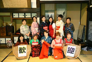 畳の部屋で色とりどりの着物やエプロンを着た女性たちが「歓迎」の文字を持って笑顔で並んでいる集合写真