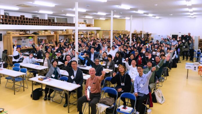 多くの参加者の皆さんが会場内の席に着き、手を振って笑顔を見せている『南三陸いのちめぐるまち学会』の集合写真