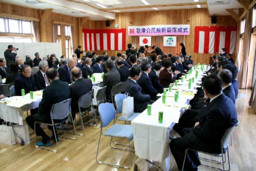 紅白の幕が飾られた会場で、壇上には「祝 歌津公民館新築落成式」の看板が掲げられ、白いテーブルクロスがかかった長いテーブルに大勢の来賓がスーツを着て着席している、式典会場の様子の写真