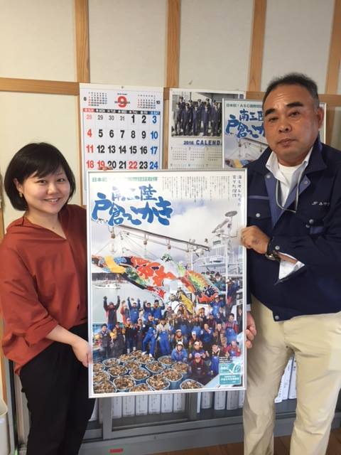 カレンダーやポスターが貼られた壁の前で、「南三陸 戸倉っこかき」と書かれた額に入ったポスターを両側から持っている女性リポーターと阿部さんの写真