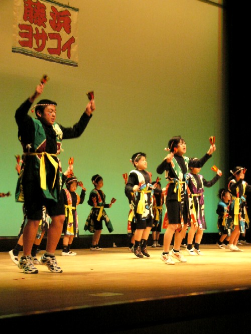 ホール内のステージ上で、子どもたちが黒い衣装に黄色と緑の帯を締め、手に鳴子を持って力強く腕を振り上げて踊りを披露している発表会の様子の写真