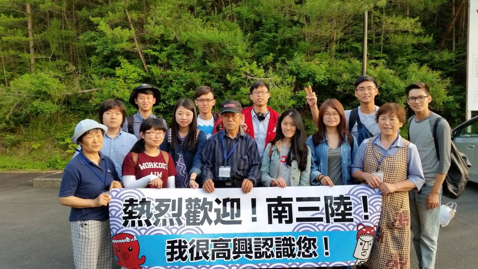 緑に囲まれた屋外の駐車場で、「熱烈歡迎！南三陸！我很高興認識您！」と中国語で書かれた横断幕を掲げ複数の人物が並んで立っている写真