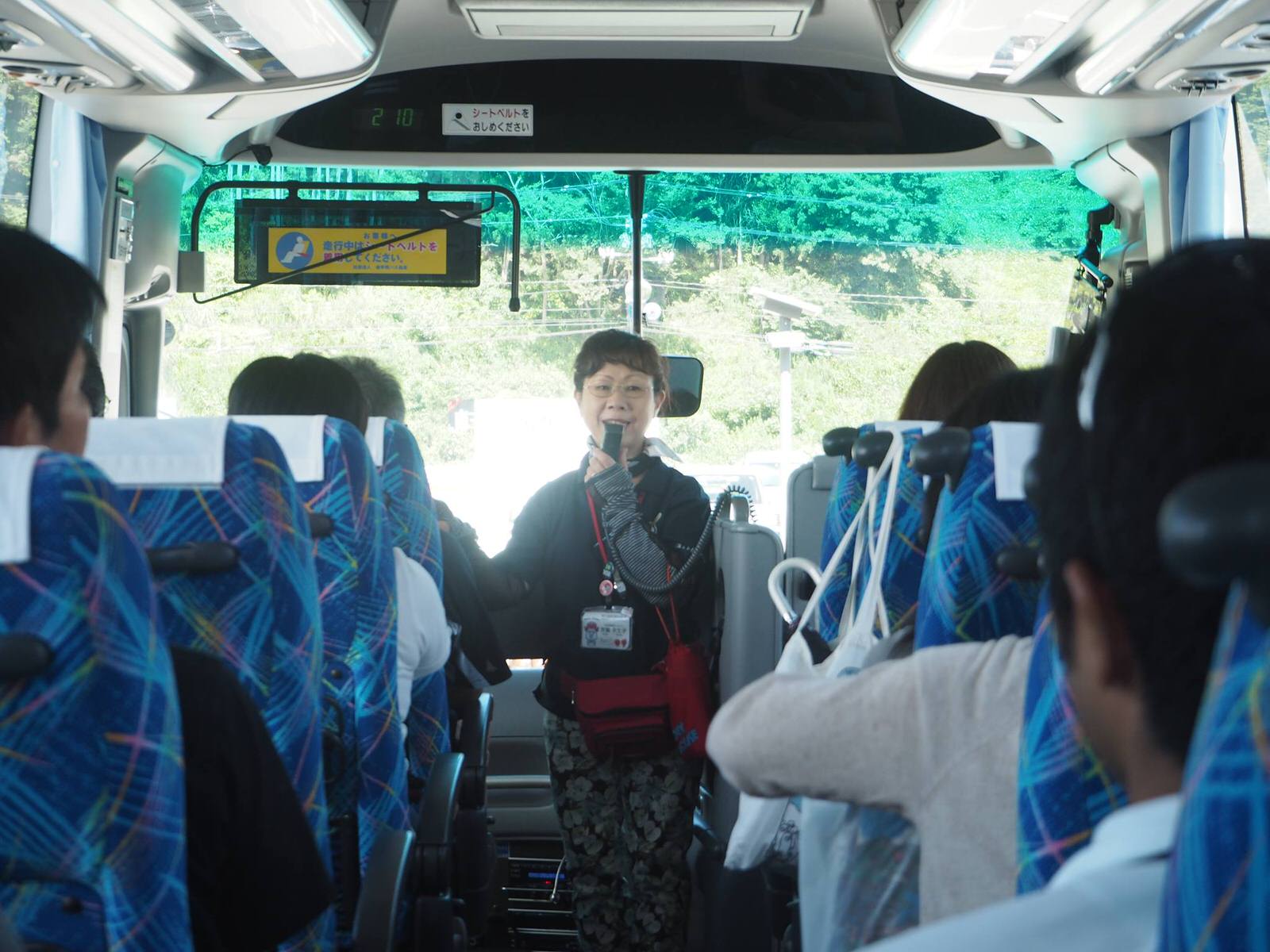 複数の乗客が座っている青い模様のシートが並ぶバスの車内で、通路の前方にマイクを持った女性が立っている語り部プログラムの様子の写真