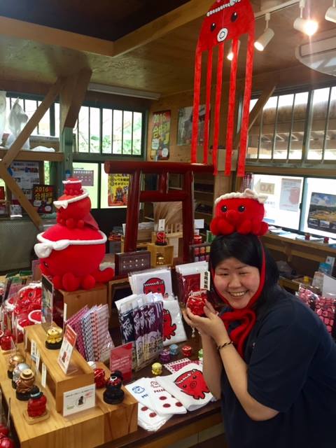ねじり鉢巻きを巻いた赤いタコのキャラクター「オクトパス君」のグッズが並ぶ棚の前で、オクトパス君の被り物を頭にかぶった女性が、グッズの一つを手に持っている写真