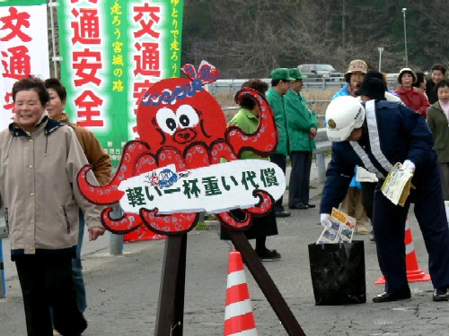 「軽い一杯重い代償」と書かれた赤いタコのマスコットの看板と交通安全ののぼり旗がたち行き交う人に飲酒運転根絶の呼びかけを行っている様子の写真