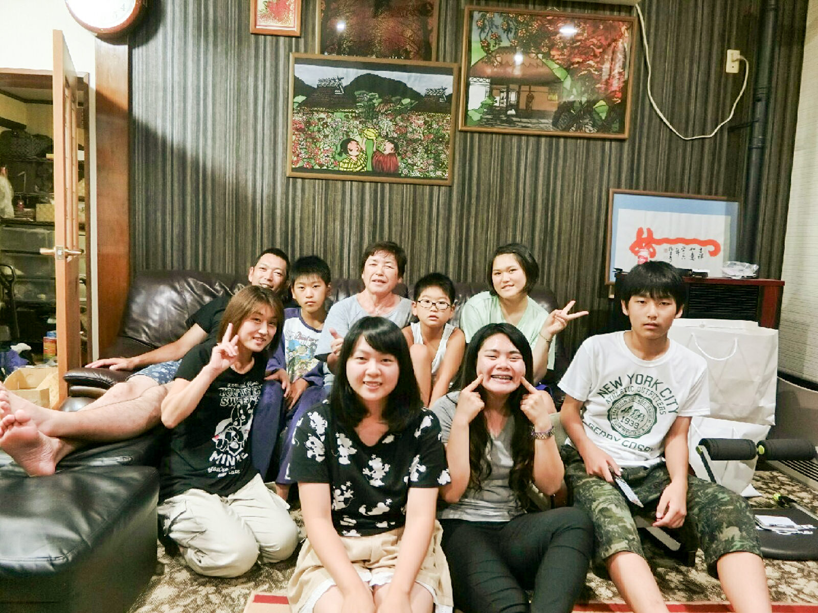 額入りのアート作品が壁に飾られた室内で、9人の人物が集まって座り、カメラに向かってポーズをとっている写真