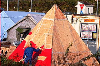 三角のピラミッド形に組み立てられた構造物に、新井さんが模様の下地として赤い色を塗っている作業の様子を写した写真