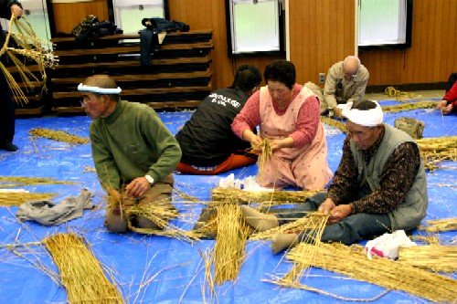 複数の関係者が青いビニールシートの上で、稲わらを用いて熱心にしめ縄づくりをしている様子の写真
