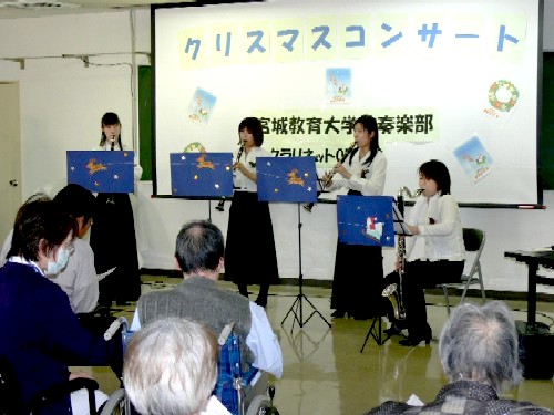 「クリスマスコンサート」と表示されたスクリーンを背景に、上下同じ色合いの白と黒の服装をした女性4人がクラリネットやバスクラリネットなどの楽器を演奏しており、手前で参加者が演奏を聞いている様子の写真