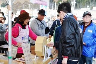 屋外のイベント会場でテーブルの上に置かれたガラガラ抽選器を男性が回している様子が写っており、その周囲ではスタッフや来場者が見守っていて、にぎやかな雰囲気が感じられる抽選会の写真