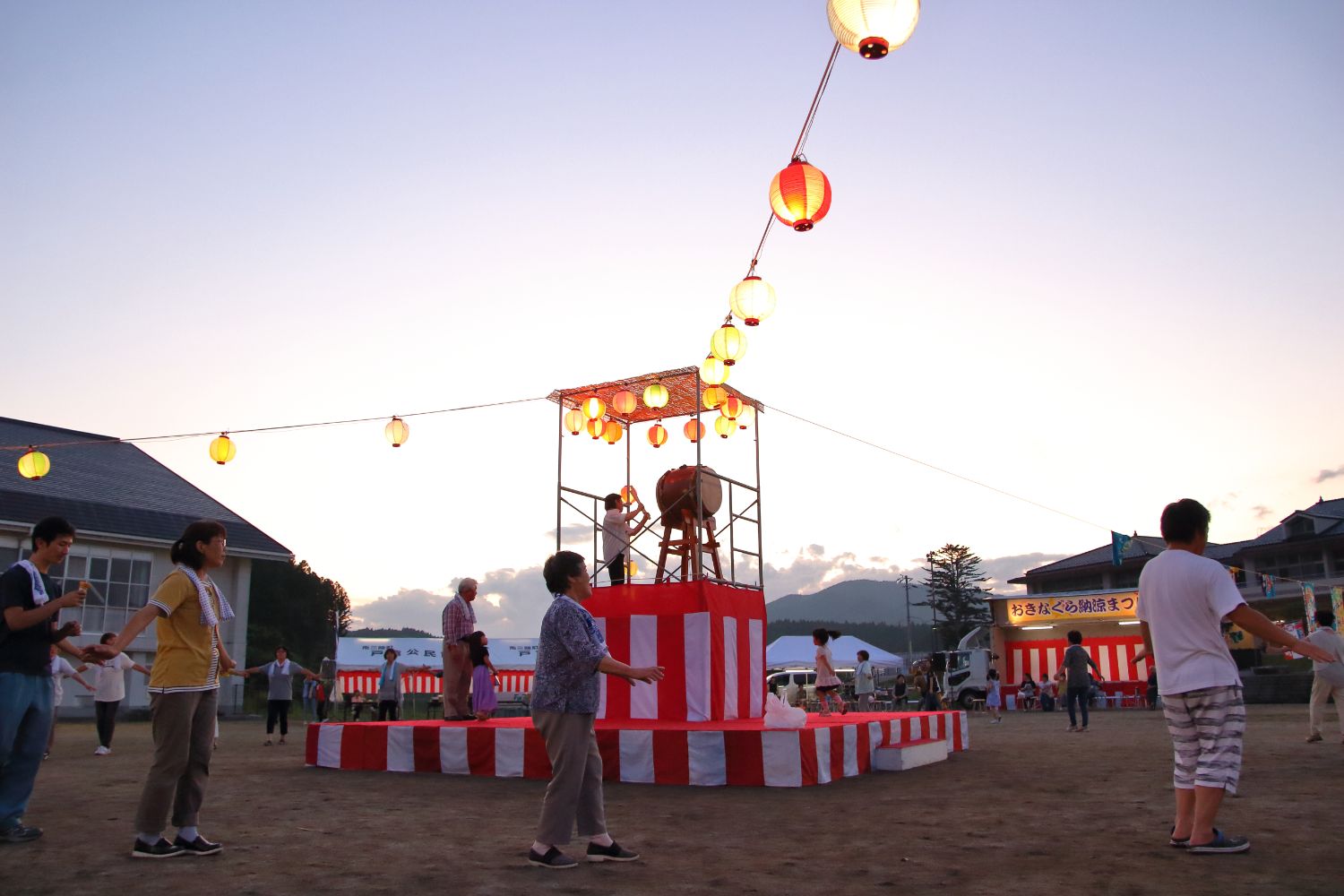 夕暮れの空の下、提灯が灯り始めたやぐら櫓の周りで、男女が輪になって盆踊りを踊っている写真