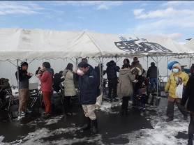 お椀を手に持った来場者たちが、雪の残る地面に立てられたテントの下に集まっている様子の写真