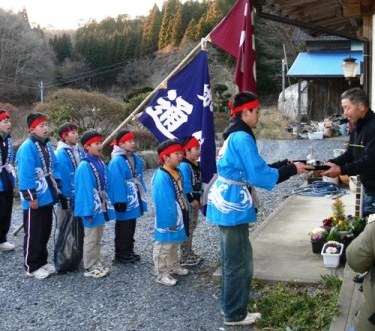 水色の法被と赤いハチマキを着用した子どもたちが、1人の子どもが青い大きな旗を掲げ、家の軒先で男性とやり取りを行っている小正月行事の写真