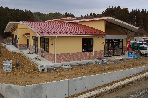 鮮やかな赤色の屋根と黄土色の外壁、茶色のレンガ風のタイルが特徴的で、周囲にはコンクリートの擁壁が見える建設途中の名足保育園の新築工事の写真