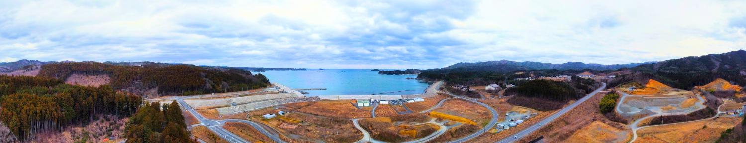 湾に面した海岸沿いの地域の中央には静かな青い海が広がり、その周囲には山と森に囲まれた造成地や曲がりくねった道路が見え、ところどころに建物や施設が点在している戸倉地区の全景の昼間の風景を高台から定点カメラで撮影した写真