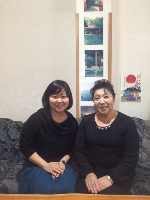 写真が飾られた壁際に置かれたソファに並んで座り、笑顔の女性リポーターと黒い洋服を着た女性の写真