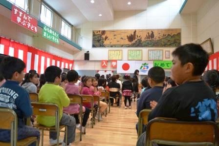 左右に紅白幕が飾られた多目的ホールに在校生の児童たちが座り、前方に新入生が立っている様子を撮影した写真