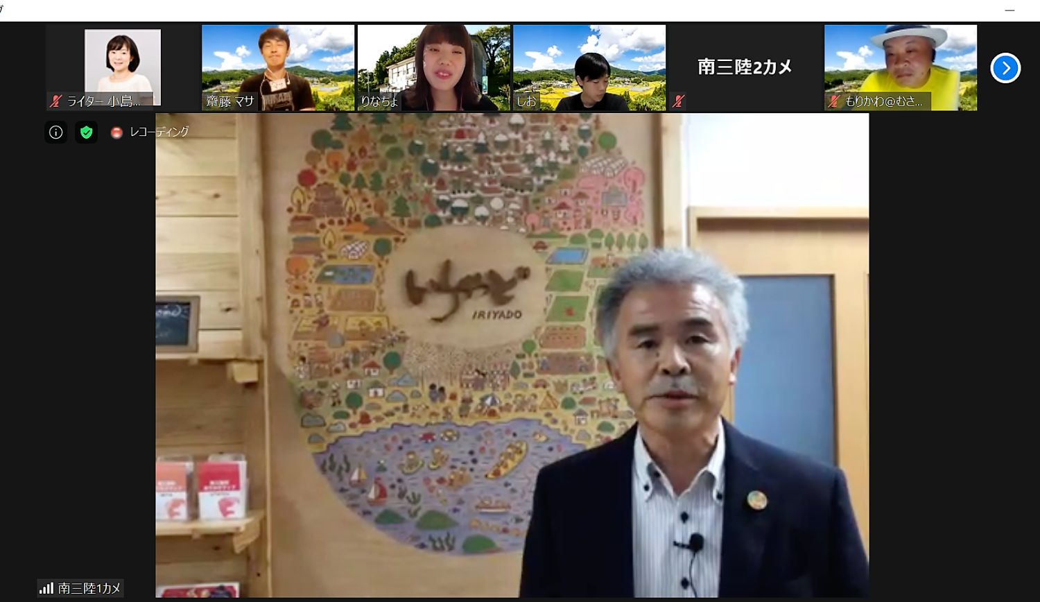 カラフルな絵や文字で「いりやど」と書かれた丸い看板の前で阿部忠義さんが話がしをしているオンラインツアーの様子を写した写真