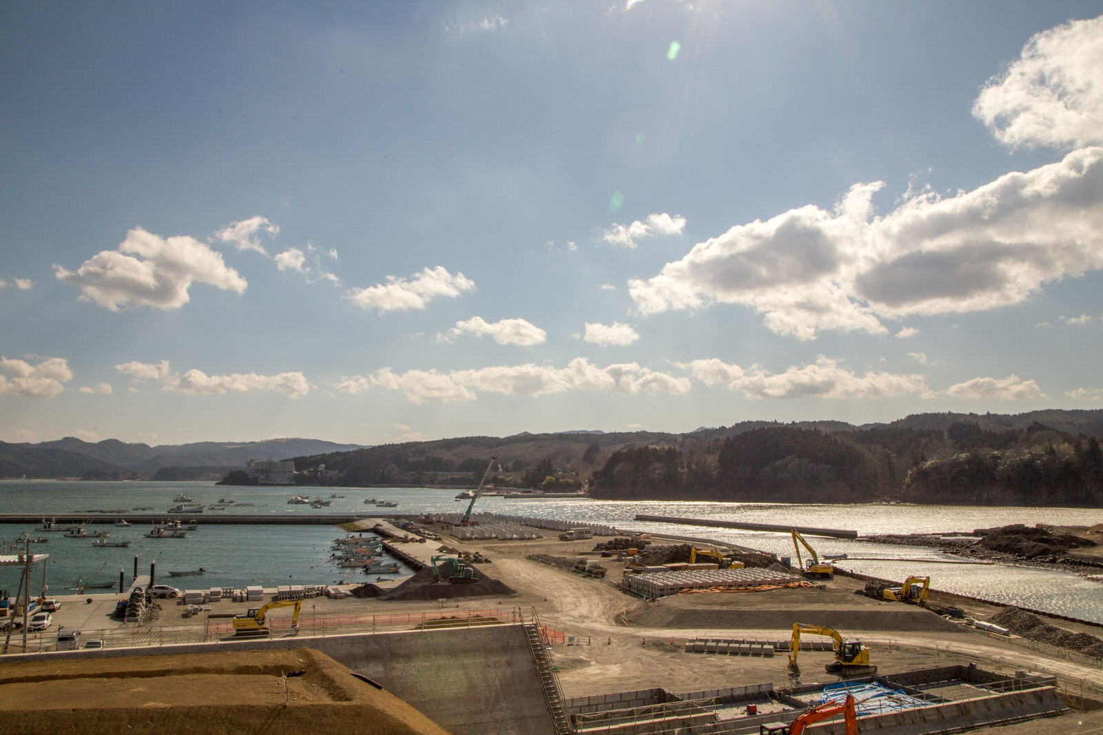 遠くに山並みが見える港湾工事が進む海沿いの場所で、クレーンや重機が作業を行っている復興中の風景の写真