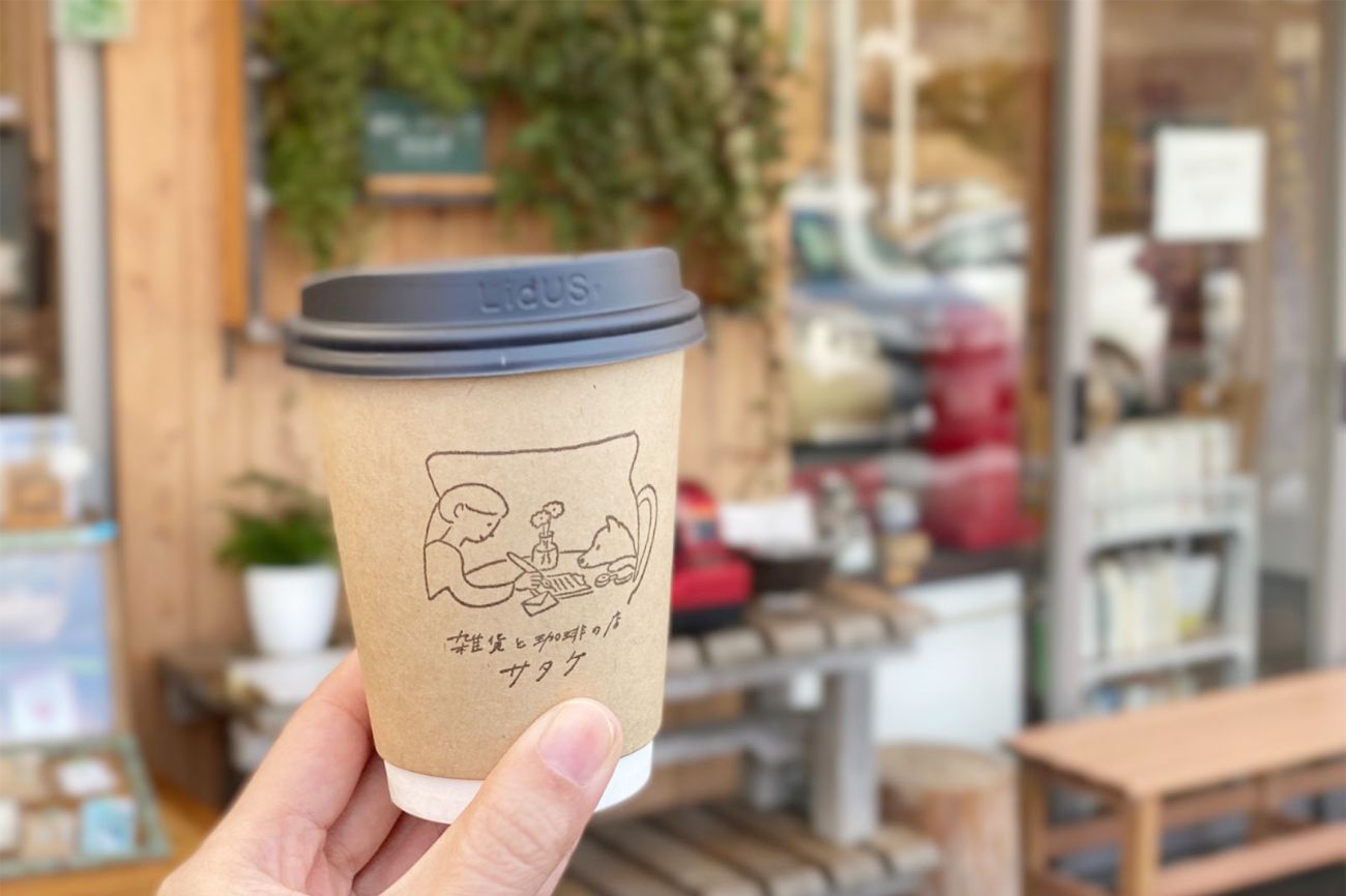 木製ベンチや観葉植物などがある店先で、「雑貨と珈琲の店サタケ」とイラストが描かれた紙コップを手に持っている写真