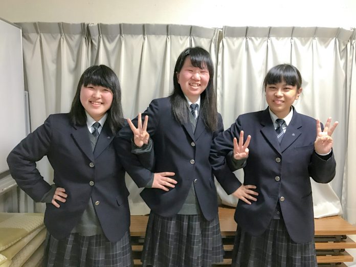 3人の制服姿の女子高校生が、左手の指で3を表し、右手を腰に当てたポーズをして笑顔で並んで立っている写真