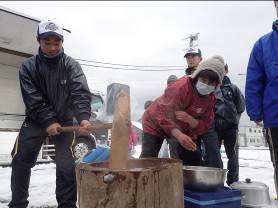 木製の臼に入った餅に男性が杵を振り下ろし、女性が合の手を入れようと準備をしてる餅つきの様子の写真