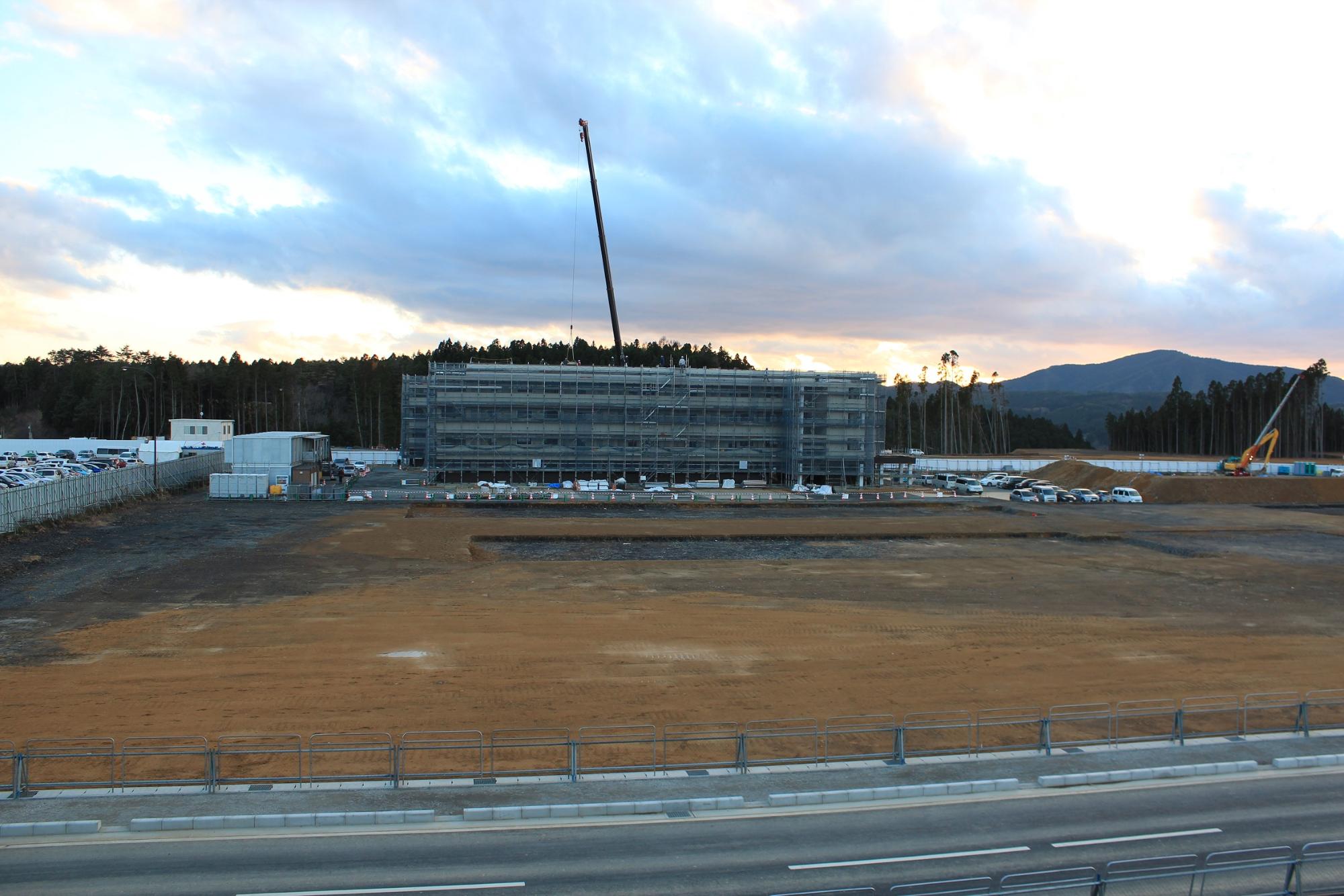 手前に整地された広大な敷地が広がり、奥には足場に覆われた建設中の建物があり、中央のクレーンがその上に高く伸びている写真