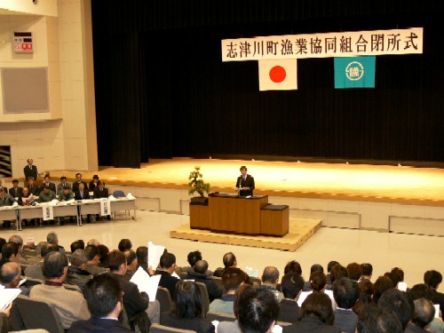 多くの参加者が着席するホールで、「志津川町漁業協同組合閉所式」と書かれた横断幕が掲げられた厳粛な雰囲気の中、スーツ姿の男性が演台の前で話をしている閉所式の様子の写真