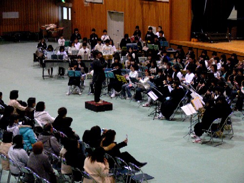 体育館で開かれた演奏会で、指揮者を中心に、生徒たちが楽器を演奏しており、観客席には多くの人々が座り、演奏を聴いてる様子の写真