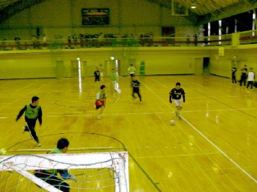 体育館で、ユニフォームを着た数人の選手たちが、ゴールポストとネットの一部が手前に写り込むアングルから、フットサルを楽しんでいる様子の写真
